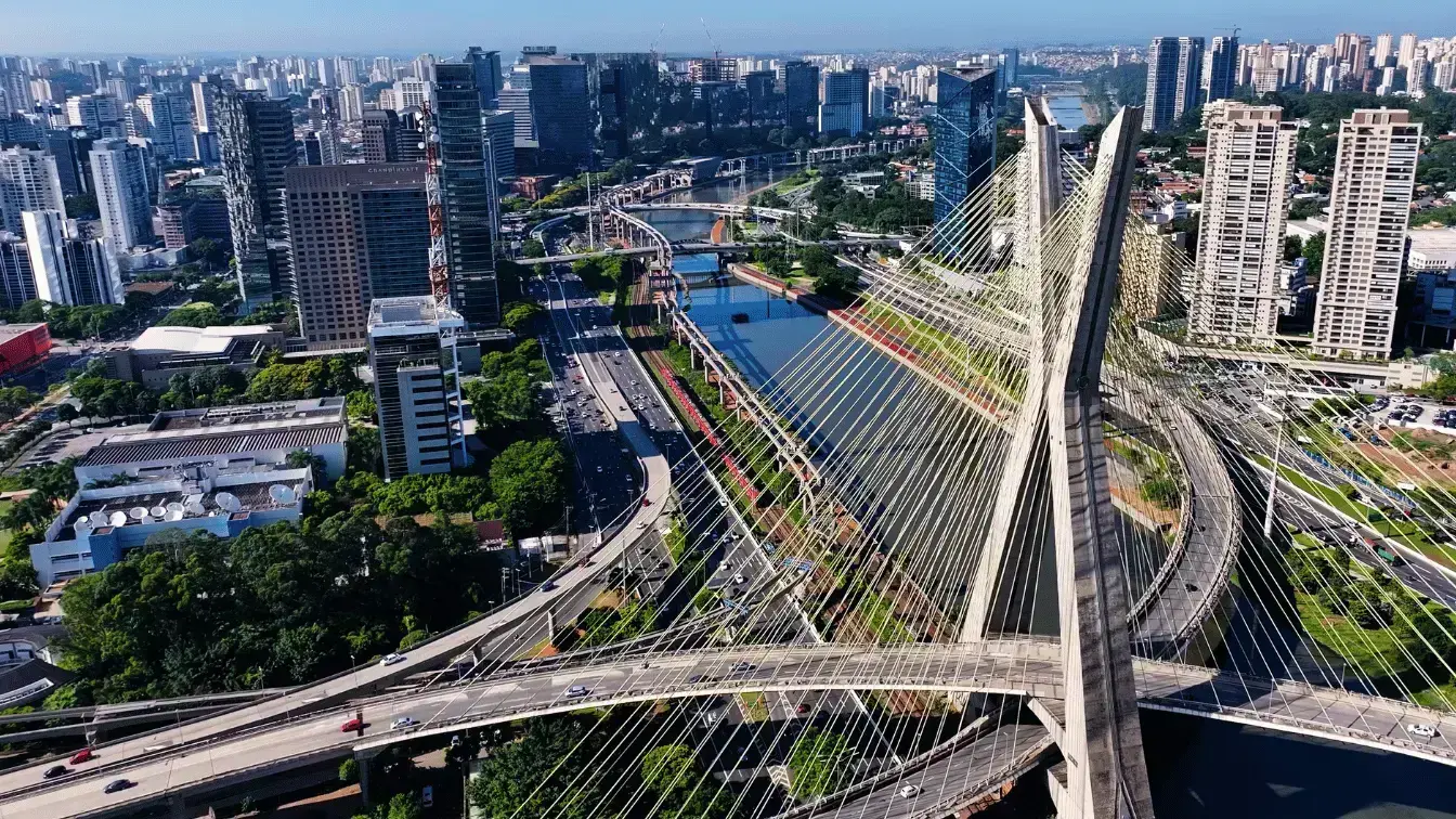 Aerial view of the Octávio Frias de Oliveira Bridge spanning the Pinheiros River in São Paulo, with dense high-rise buildings, multilane highways, and light traffic under clear blue skies.