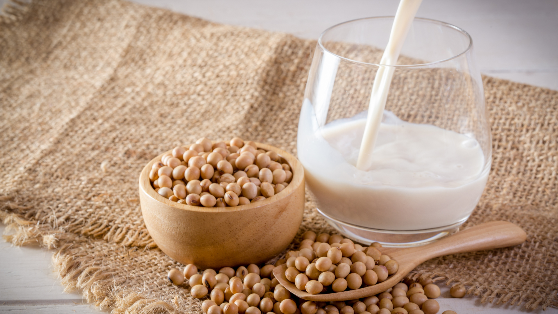 Glass of soy milk being poured beside a bowl of soybeans on a rustic burlap surface.