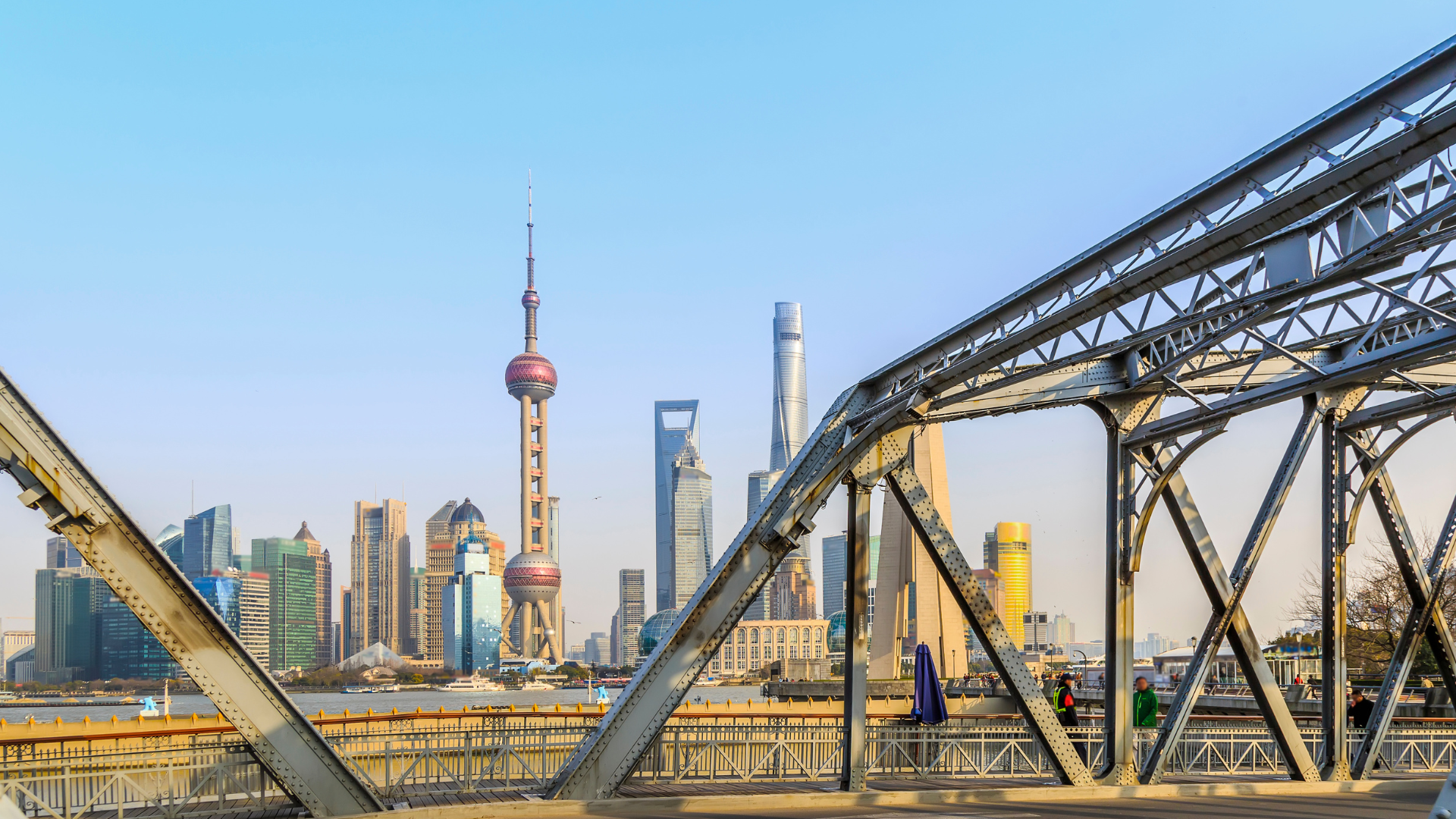 Shanghai skyline featuring the Oriental Pearl Tower and Lujiazui financial district, viewed through a steel bridge over the Huangpu River.