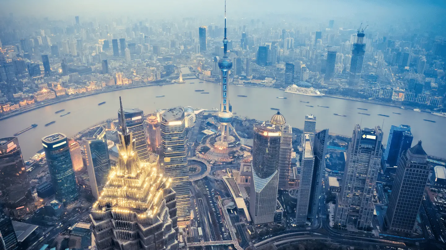 Aerial view of Shanghai’s Lujiazui financial district at dusk, featuring the Oriental Pearl Tower, skyscrapers, and the Huangpu River with boats crossing through the city skyline.